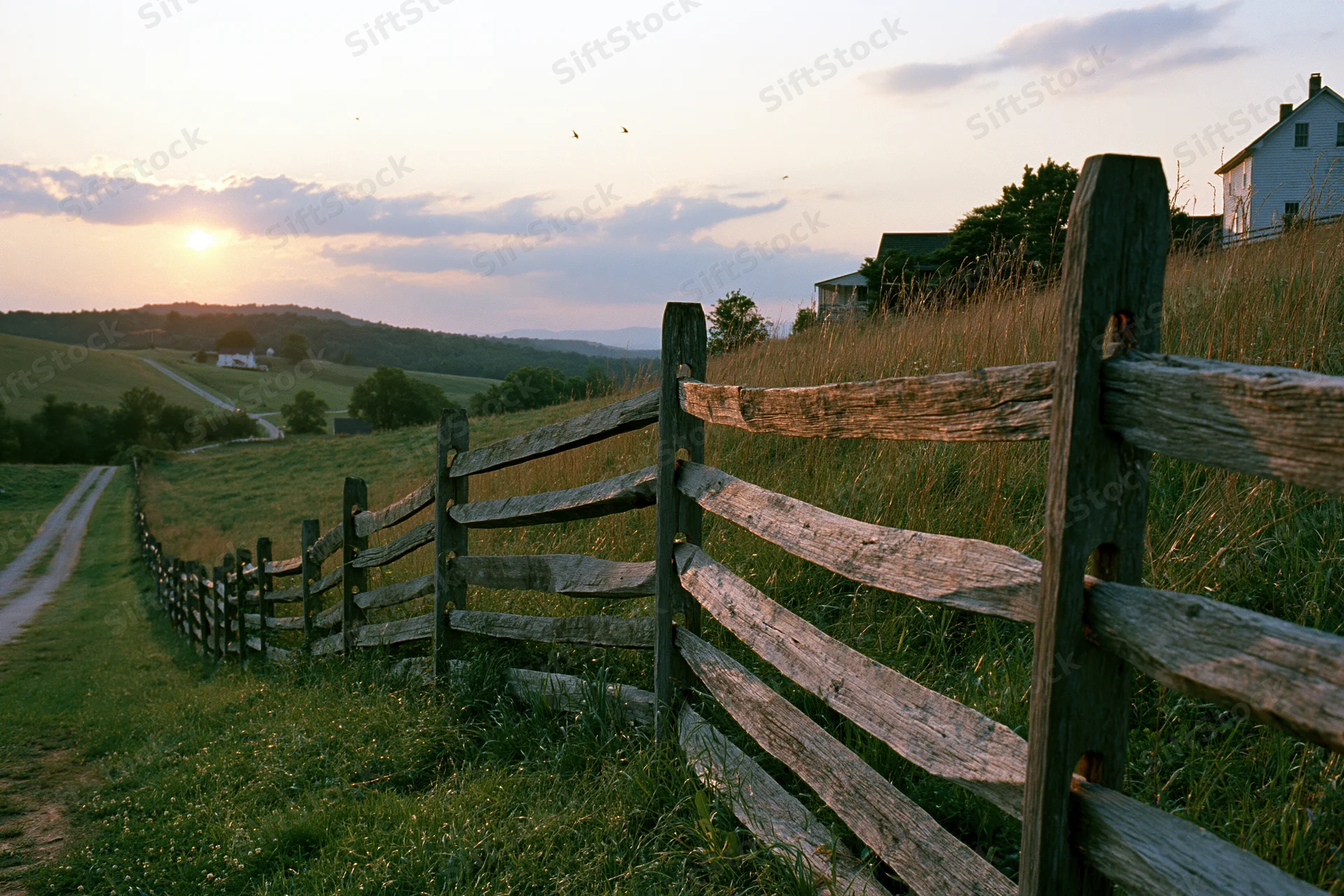 Weathered split-rail wooden fence running diagonally through a grassy field at sunset near a farmhouse