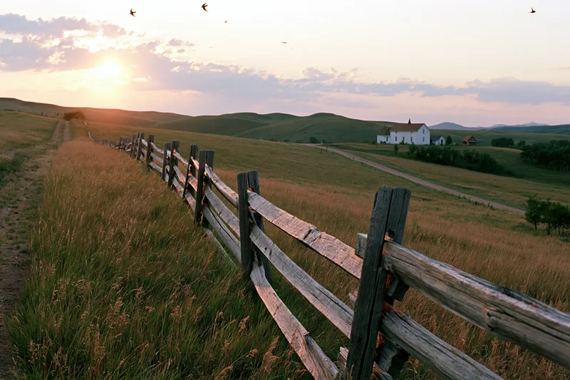Weathered split-rail wooden fence running diagonally through a grassy field toward a white farmhouse at sunset