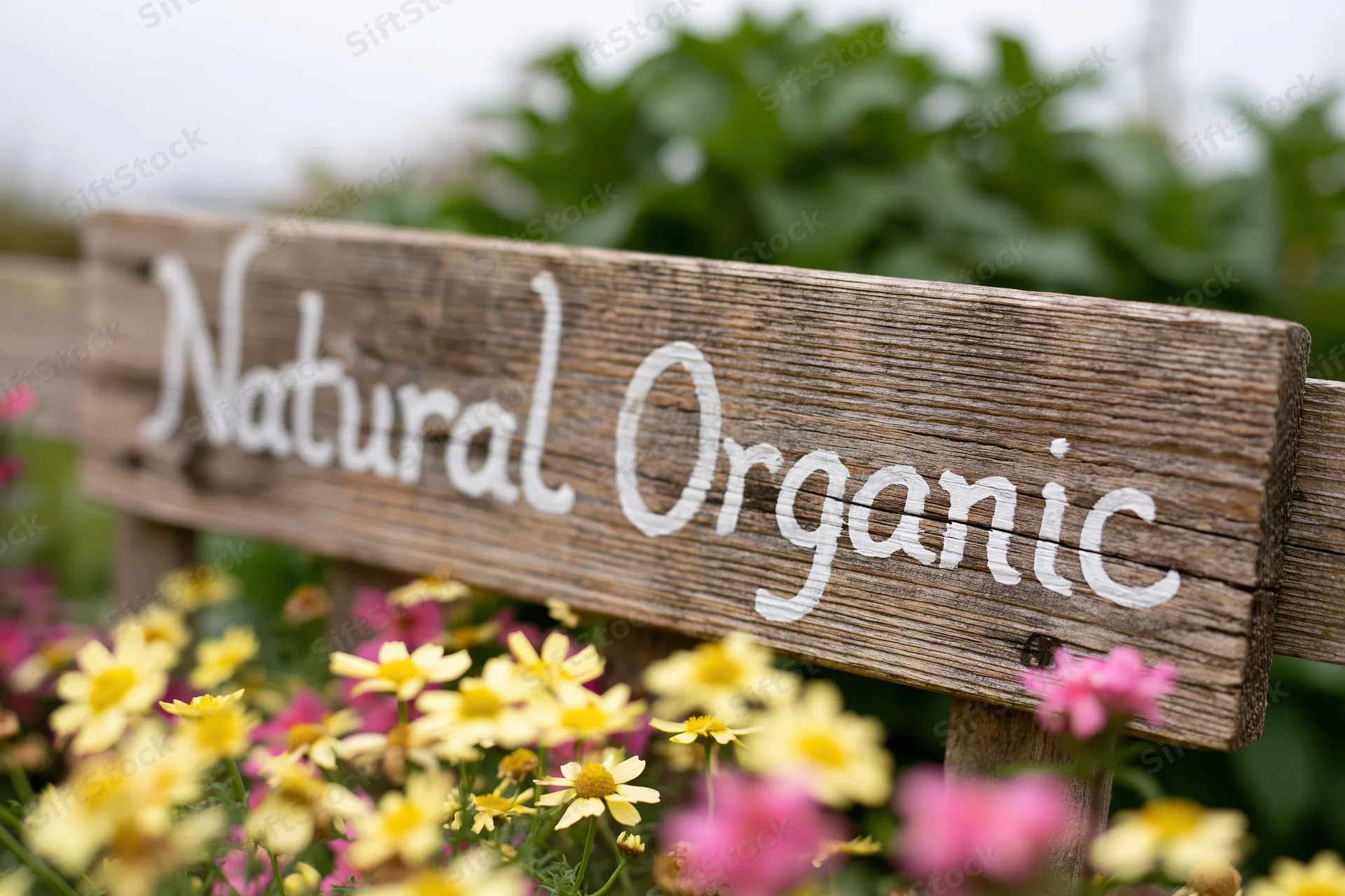 Weathered wooden sign with hand-painted white script reading natural organic in a garden with yellow and pink flowers