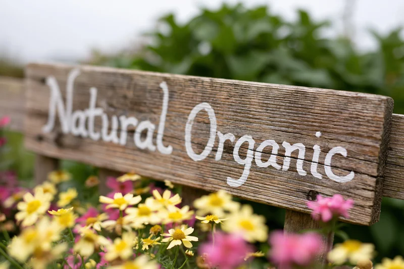 Weathered wooden sign with hand-painted white script reading natural organic in a garden with yellow and pink flowers