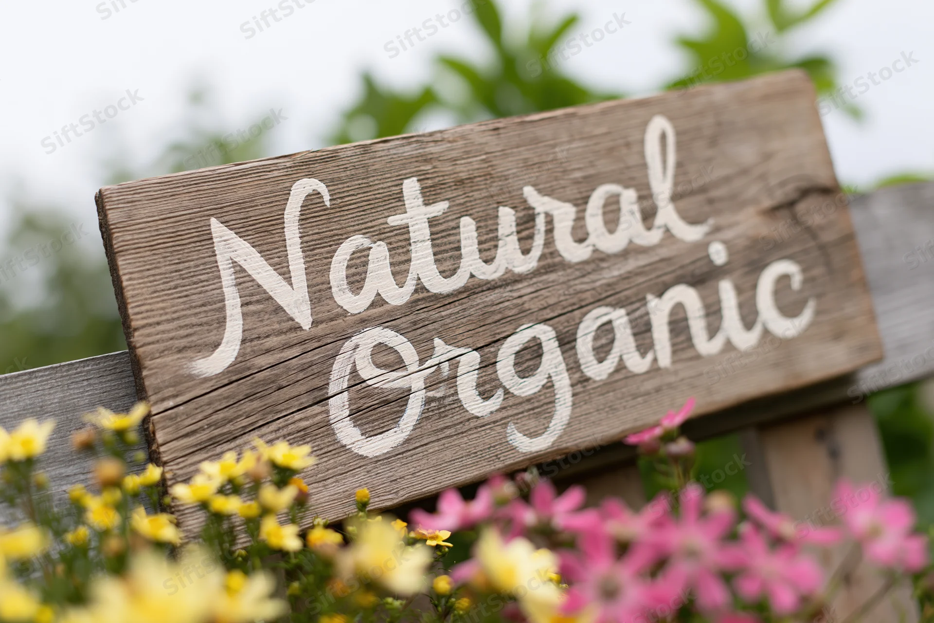 Weathered wooden sign with hand-painted white script reading natural organic in a garden with yellow and pink flowers