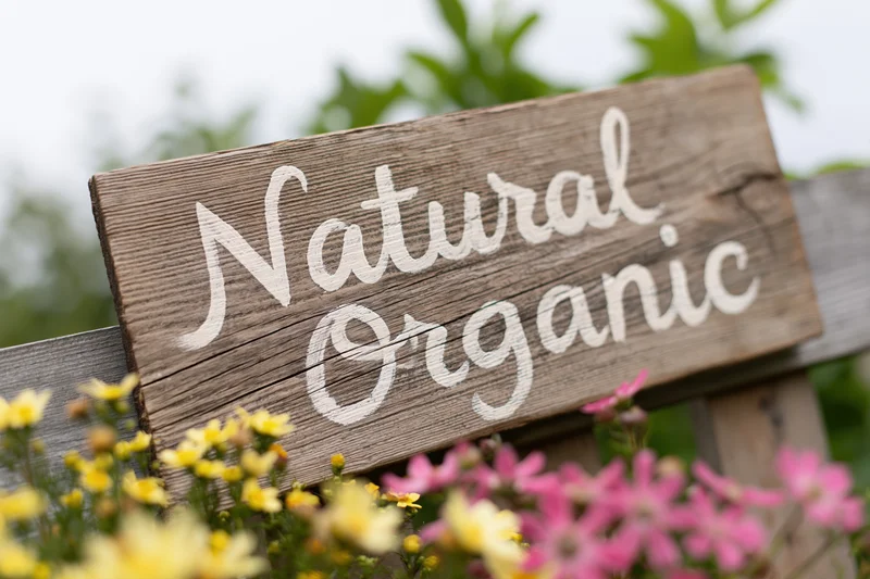 Weathered wooden sign with hand-painted white script reading natural organic in a garden with yellow and pink flowers