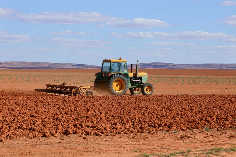 Weathered yellow-green tractor pulling a disc harrow across a red soil field under a partly cloudy sky