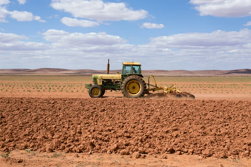 Yellow-green tractor pulling disc harrow across red soil field under blue sky with clouds