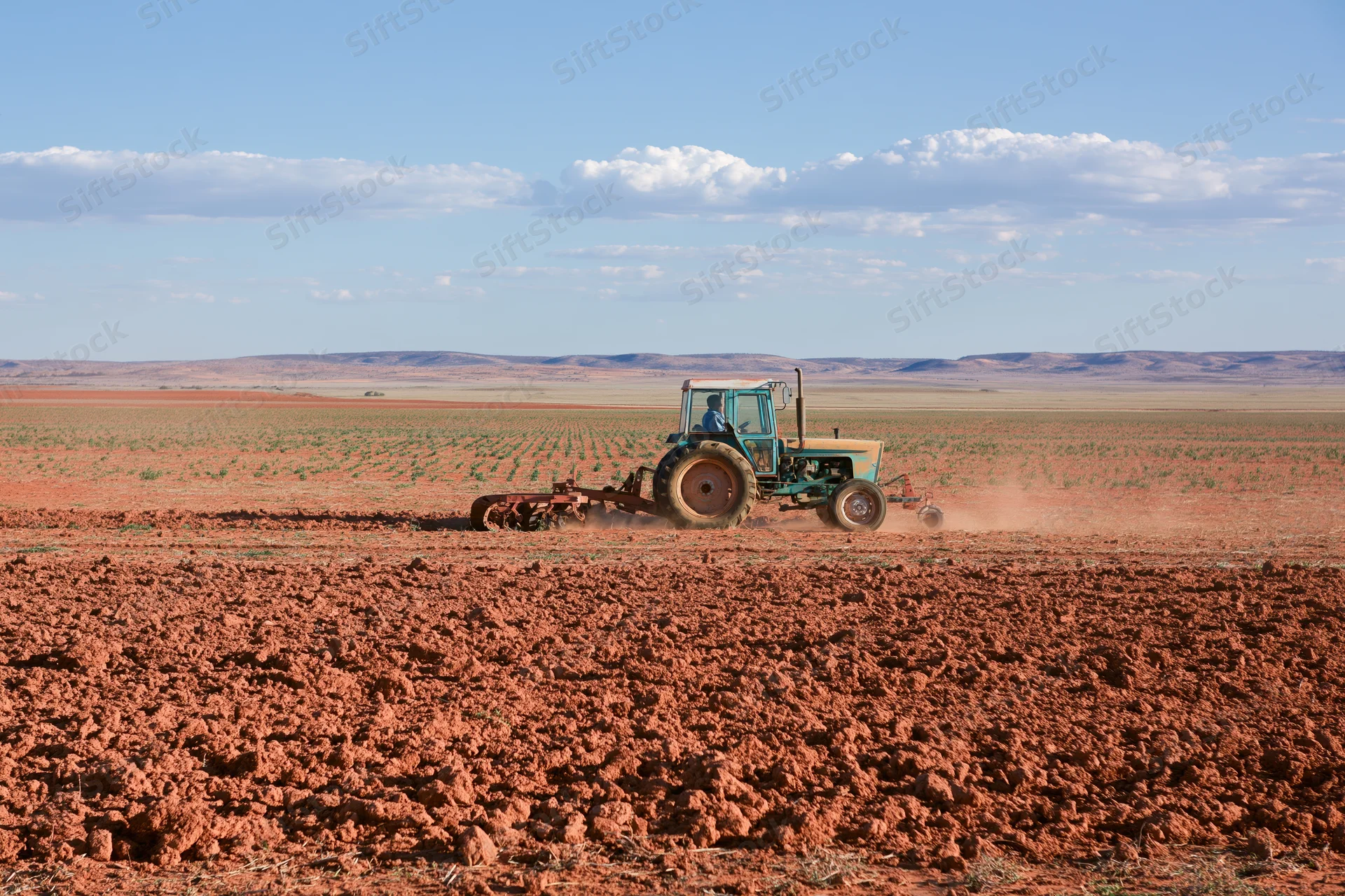 Tractor pulling disc harrow across red soil field under blue sky with clouds