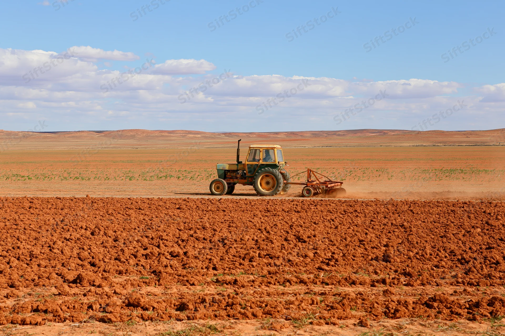 Weathered yellow-green tractor pulling a disc harrow across a red dirt field under a partly cloudy sky