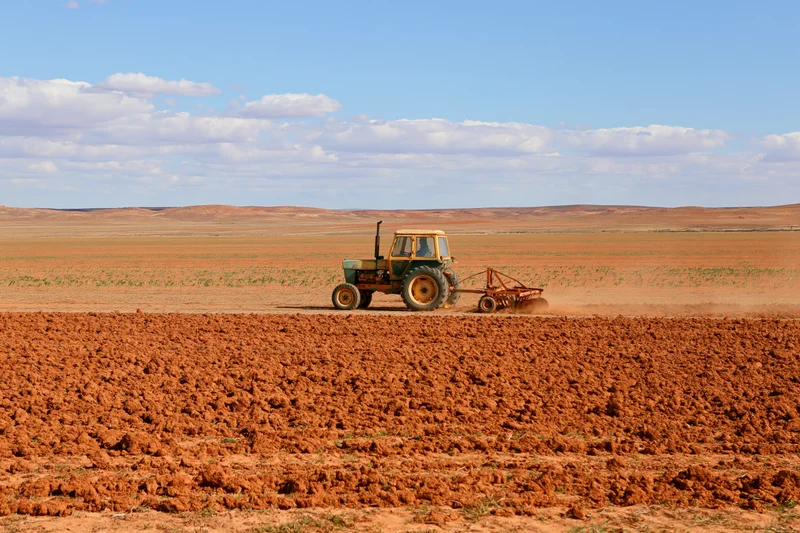 Weathered yellow-green tractor pulling a disc harrow across a red dirt field under a partly cloudy sky