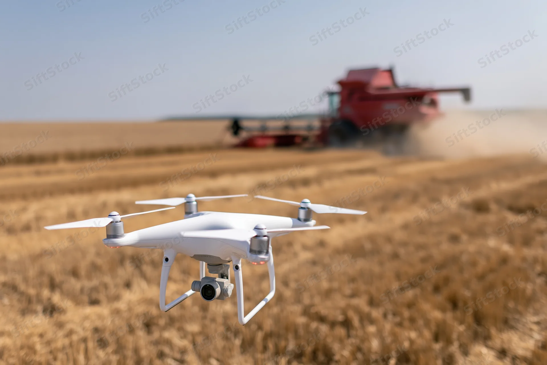 White quadcopter drone hovering low over harvested wheat field with red combine harvester in background
