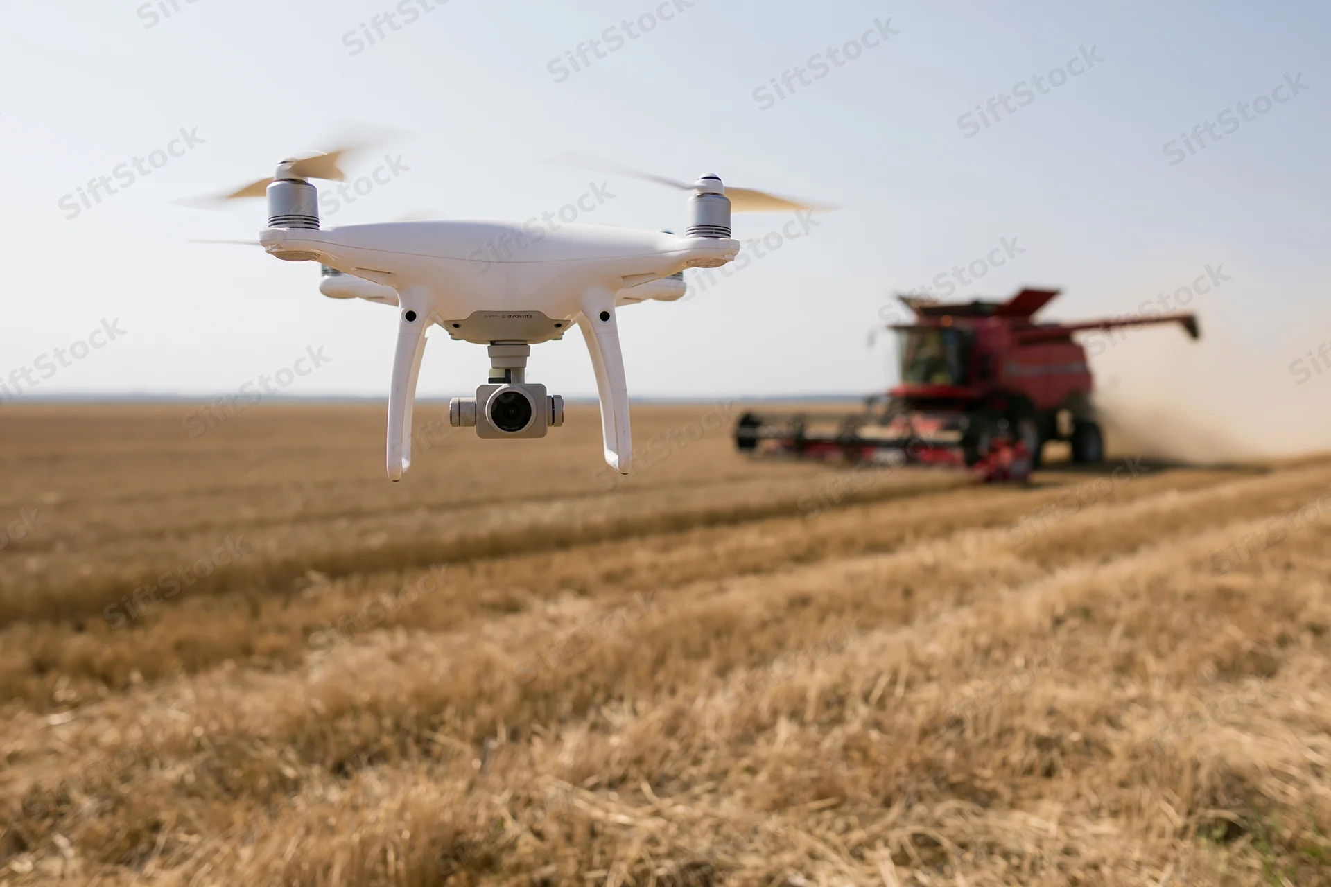 White quadcopter drone hovering low over harvested field with red combine harvester in background