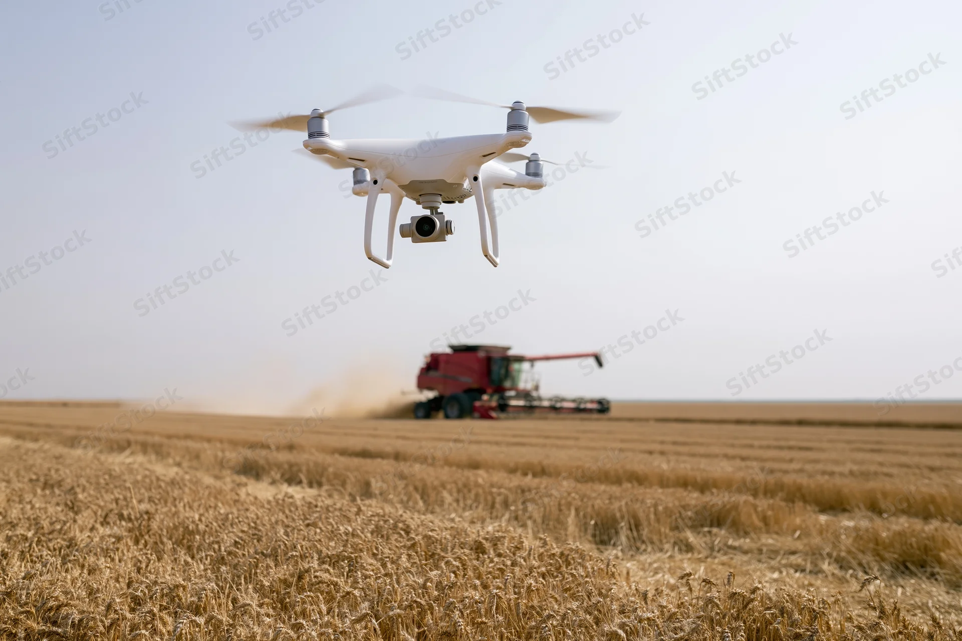 White drone hovering low over harvested wheat field with combine harvester in background