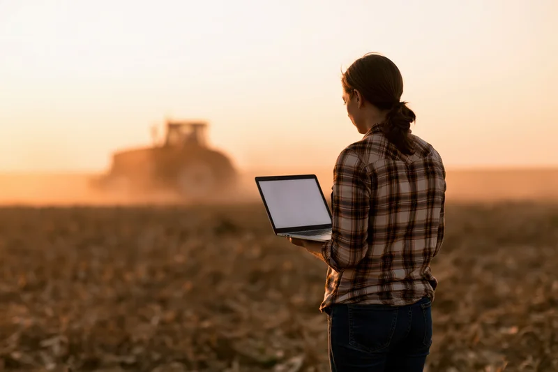 Woman holding open laptop in harvested corn field at sunset with tractor in background