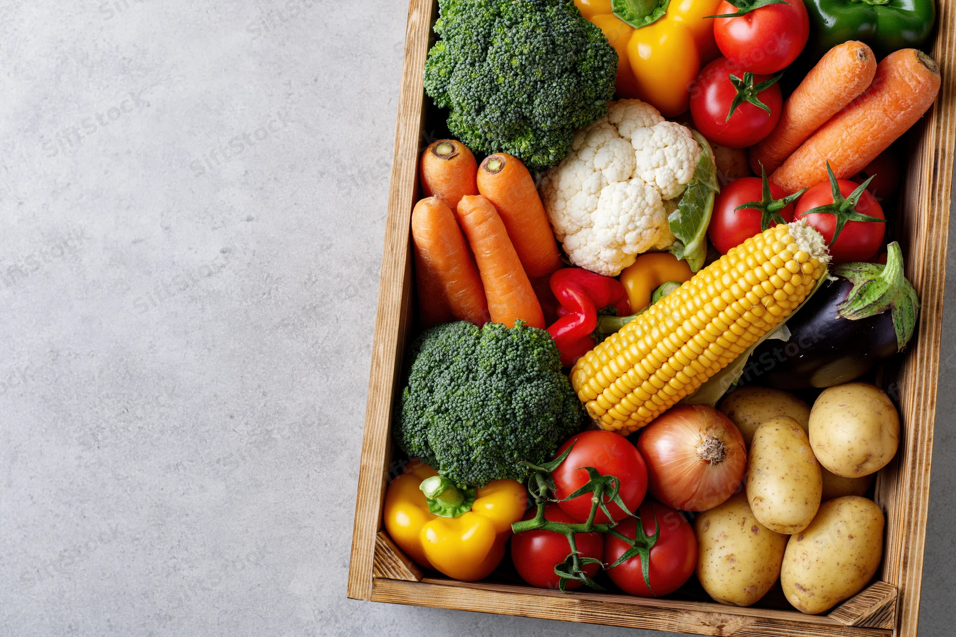 Wooden crate filled with assorted fresh vegetables arranged on a light surface with copyspace on the left