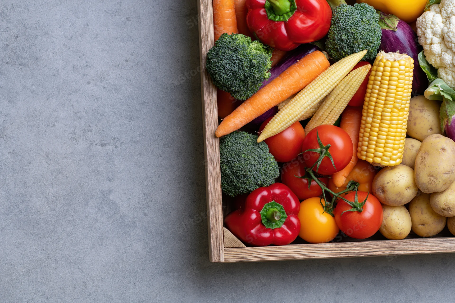 Wooden crate filled with fresh mixed vegetables arranged on gray surface with copyspace