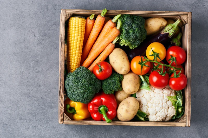 Wooden crate filled with fresh mixed vegetables arranged on gray surface