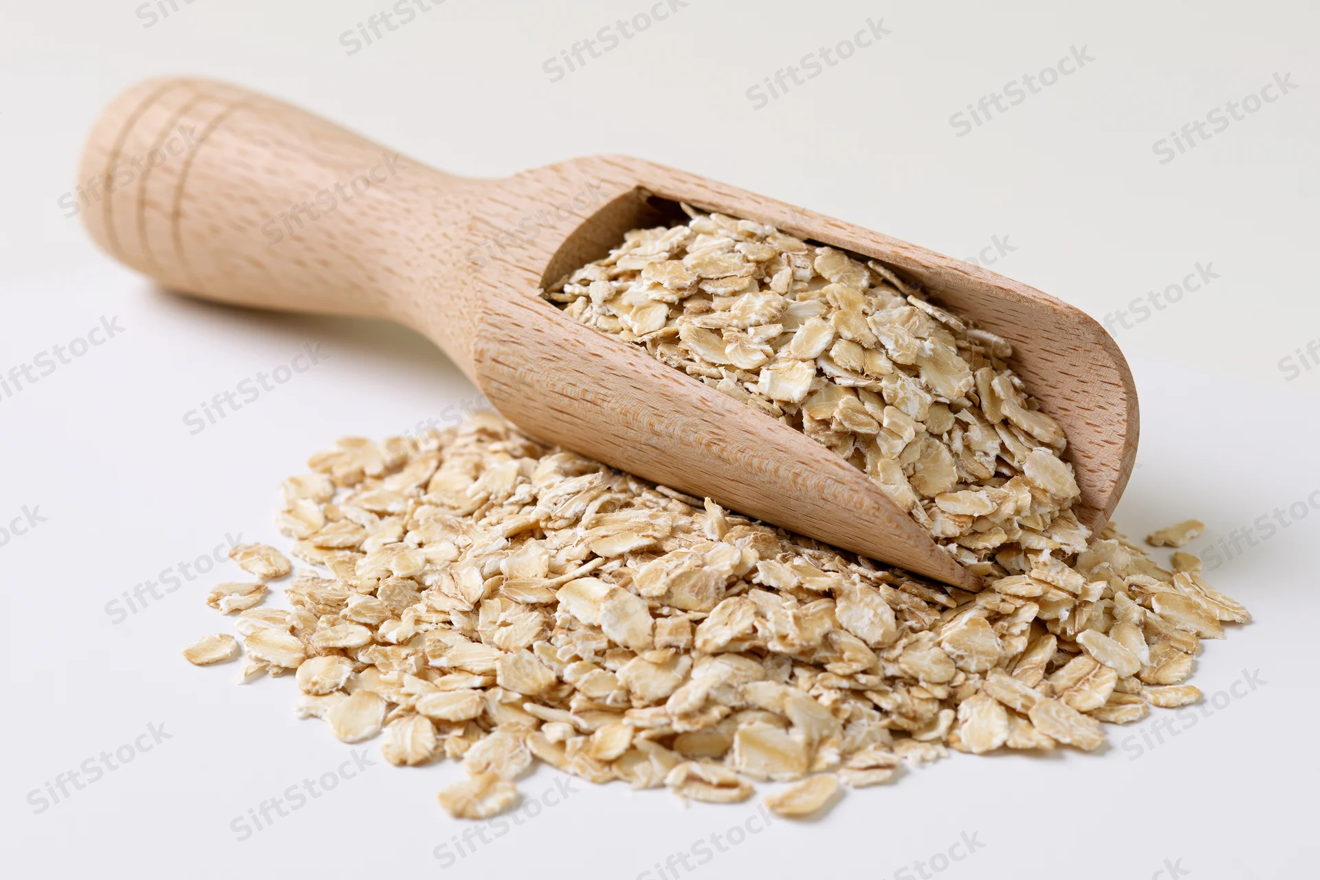 Wooden scoop resting on a pile of dry rolled oats on white surface