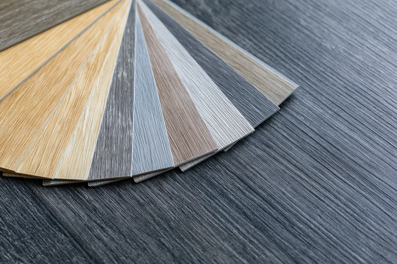 Fan of wood-grain vinyl flooring samples arranged on a dark gray textured floor