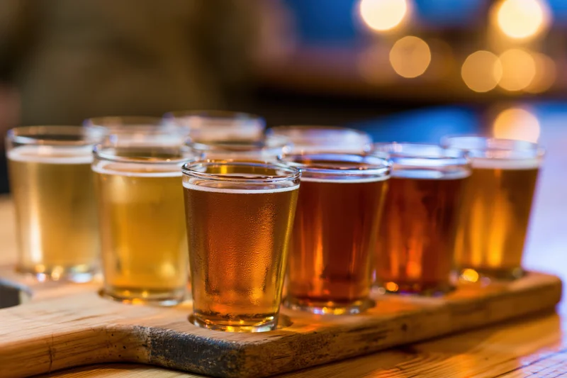 Eight small beer glasses arranged in two rows on a wooden tray with condensation and foam, set against warm bokeh lighting in a bar setting