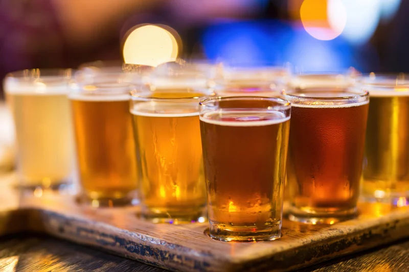 Eight small beer glasses arranged in two rows on a wooden tray with condensation and foam, in a dimly lit bar setting