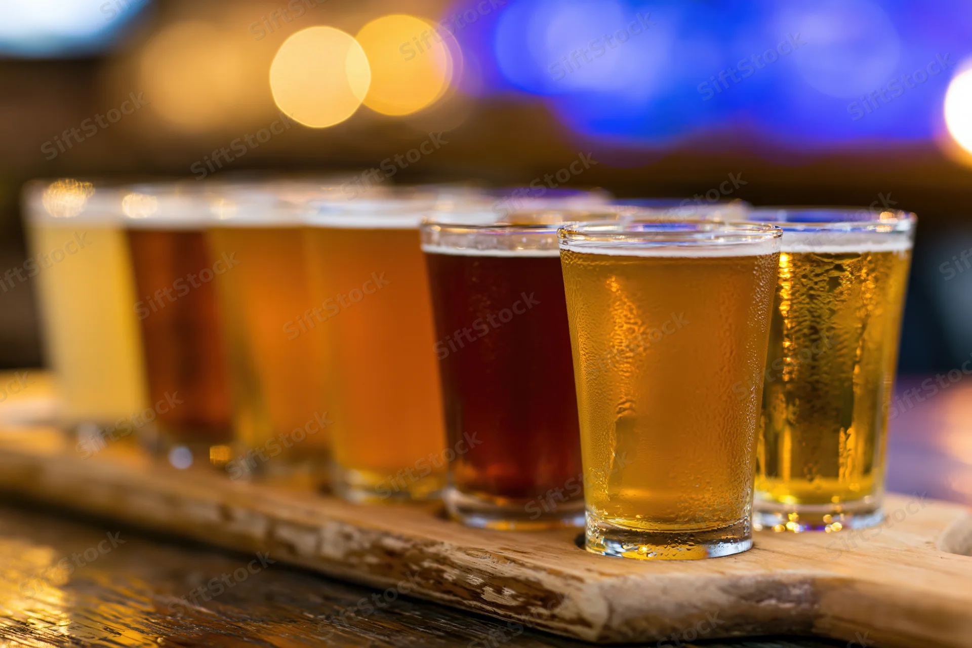 Eight small beer glasses arranged in two rows on a wooden tray with condensation, set on a bar surface under warm ambient lighting