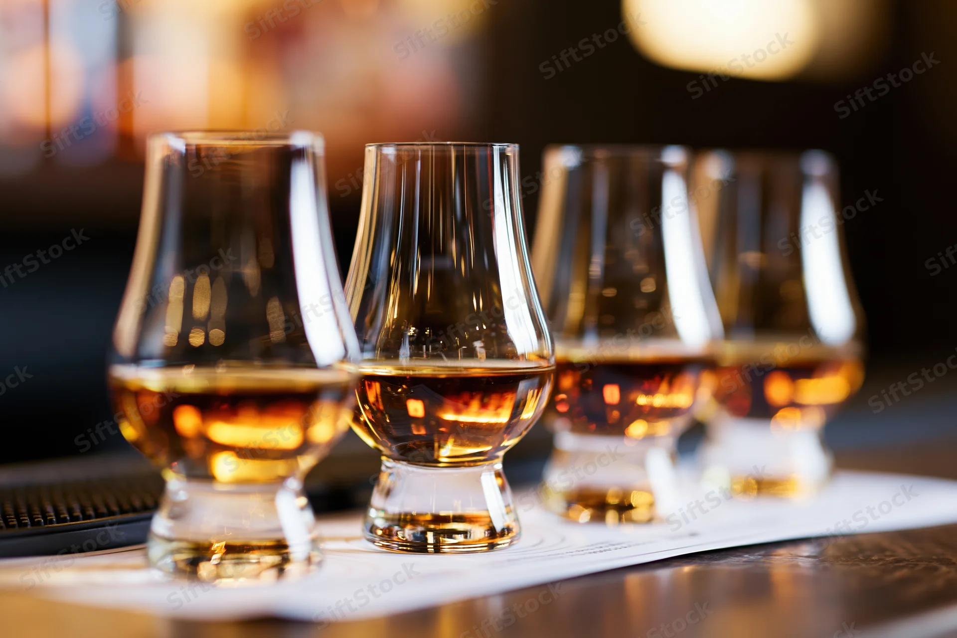 Five glencairn glasses filled with amber whisky arranged in a row on a bar top with a paper tasting sheet beneath them
