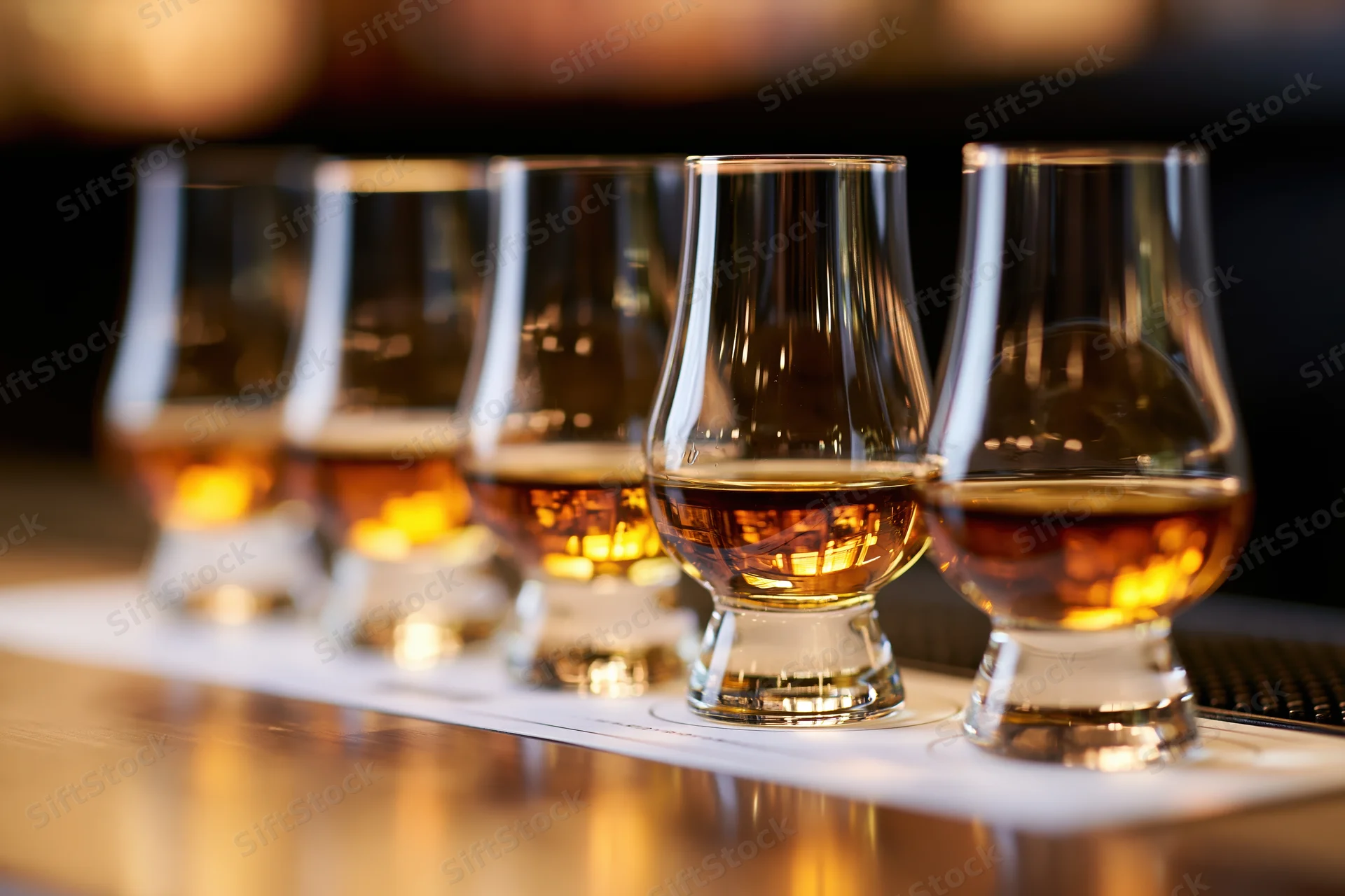 Five glencairn glasses filled with amber whisky arranged in a row on a bar mat