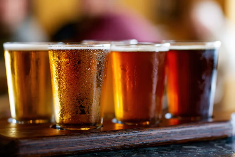Craft beer tasting flight of five glasses arranged on wooden tray with condensation and froth, in bar setting