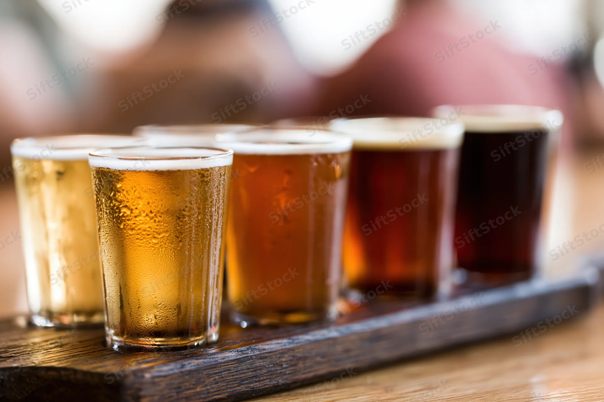 Five small craft beer tasting glasses arranged in a row on a wooden tray with condensation and foam, placed on a table in a bar setting