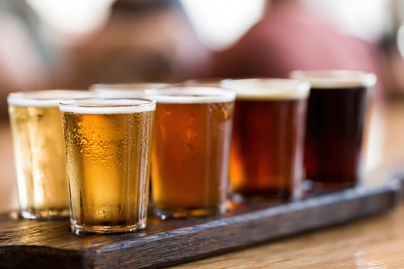 Five small craft beer tasting glasses arranged in a row on a wooden tray with condensation and foam, placed on a table in a bar setting
