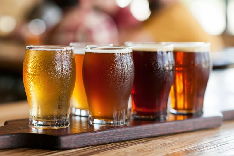 Five small craft beer tasting glasses arranged in a row on a wooden tray with condensation, placed on a wooden table in a bar setting