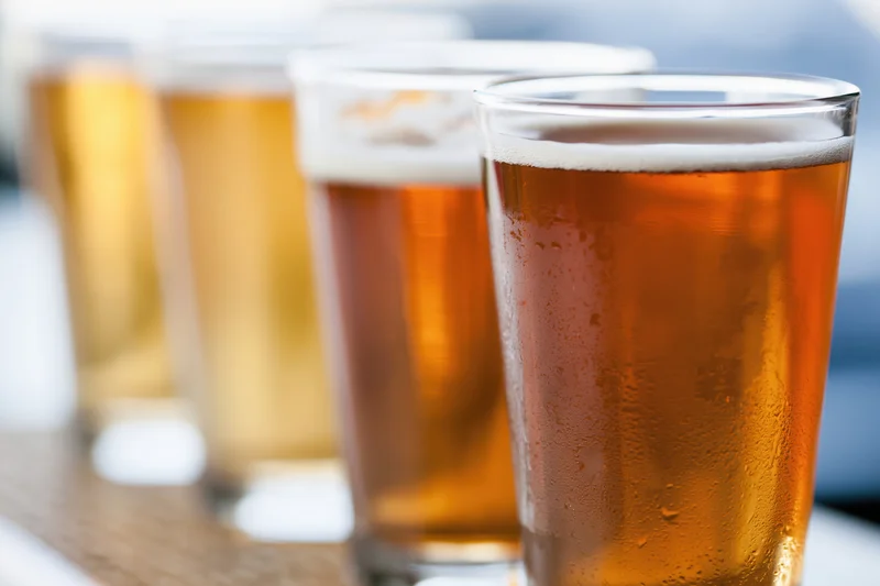 Four craft beer tasting glasses lined up in a row on a wooden surface with condensation and foam, showing amber and golden hues in shallow focus