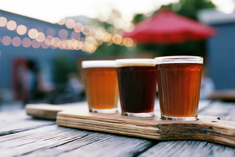 Craft beer flight in small glasses arranged on wooden tray on outdoor patio table at evening