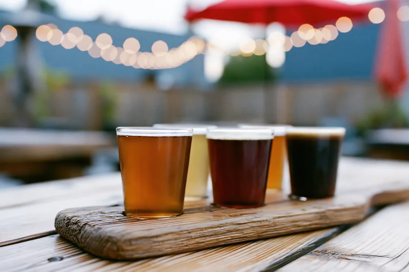 Four craft beers in small glasses arranged on a wooden tray on an outdoor patio table at twilight