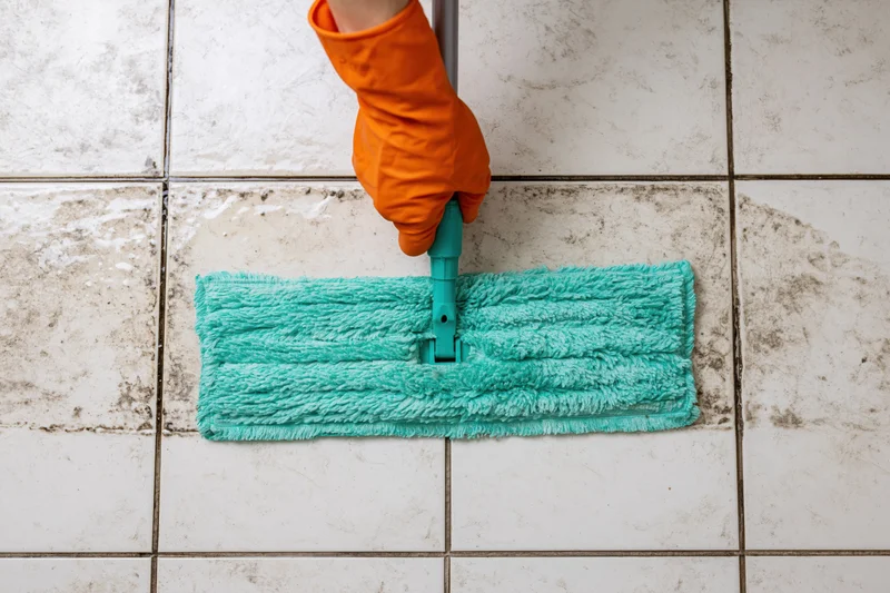 Gloved hand pushing turquoise flat mop across dirty ceramic tile floor