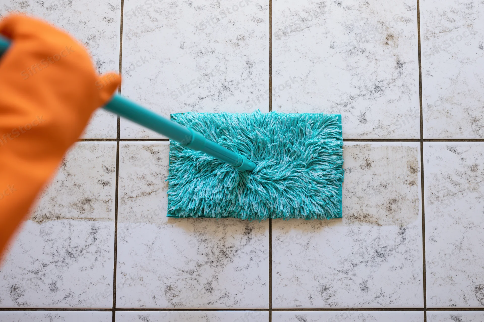 Gloved hand pushing turquoise flat mop across dirty ceramic tile floor