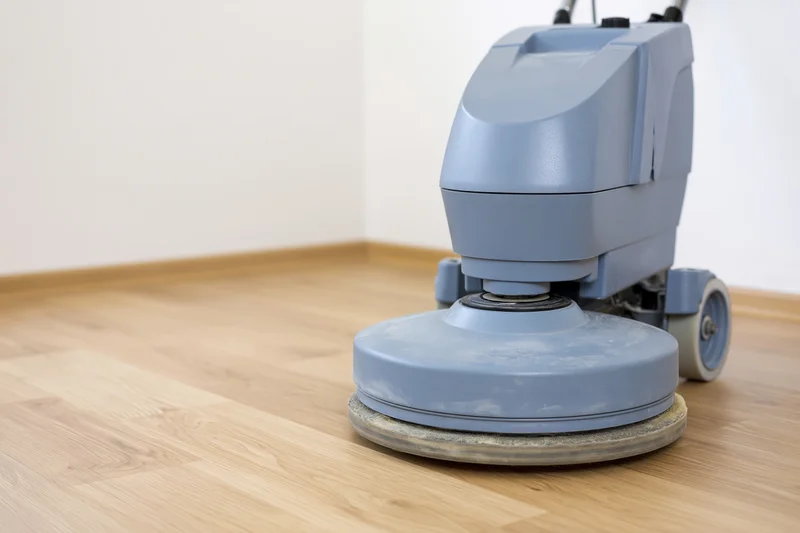 Gray-blue commercial floor polishing machine resting on light hardwood flooring in an interior room corner
