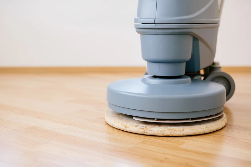 Gray-blue commercial floor polishing machine resting on light wooden floor with polishing pad attached