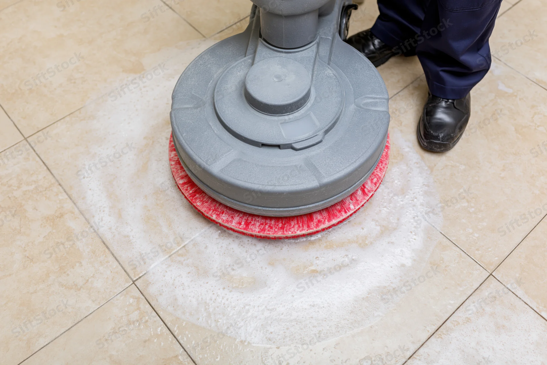 Janitor operating rotary floor scrubbing machine on tiled floor with foam and red pad