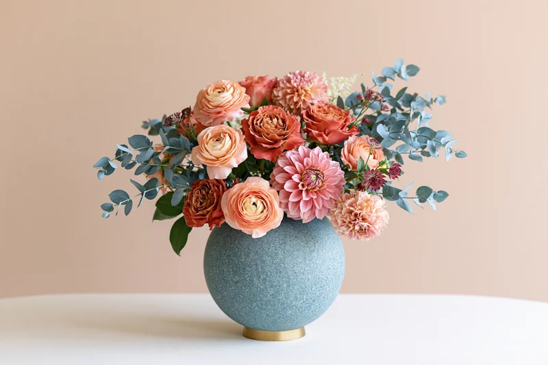 Mixed floral arrangement with peach ranunculus and rust garden roses in a blue ceramic vase on a white table