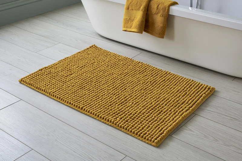 Mustard yellow chenille bath mat with raised bobble texture placed on light gray tiled bathroom floor beside white bathtub