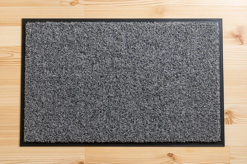 Grey doormat with black rubber border lying flat on light wooden flooring