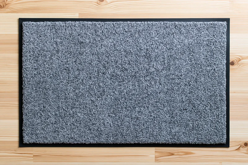 Grey rectangular doormat with black rubber border lying on light wood flooring