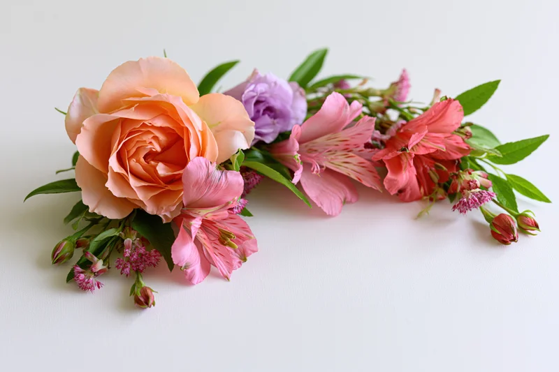 Curved floral arrangement with rose, alstroemeria, peony and greenery lying flat on white surface