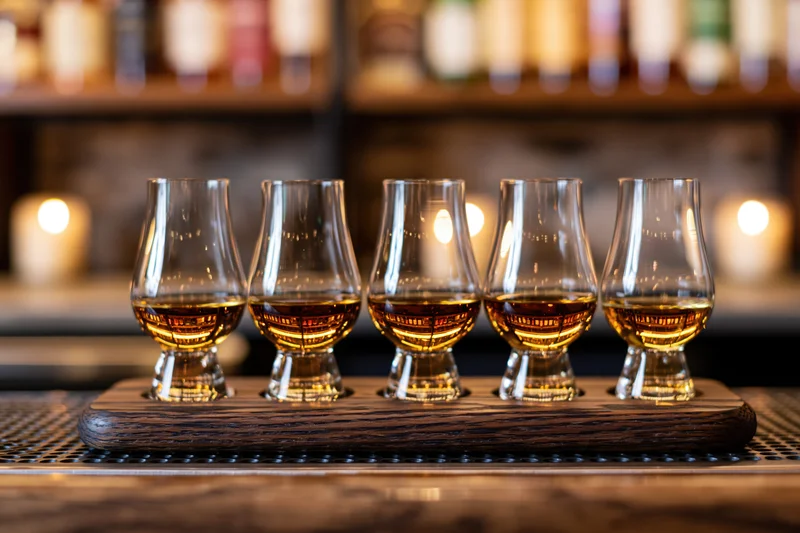 Five glencairn glasses filled with whisky arranged in a row on a wooden tray at a bar