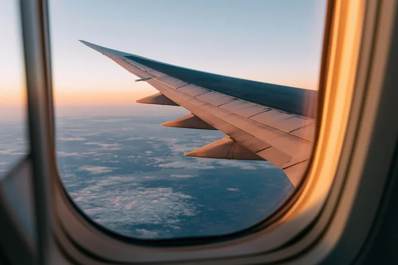 Airplane wing visible through passenger window during flight above clouds at sunset