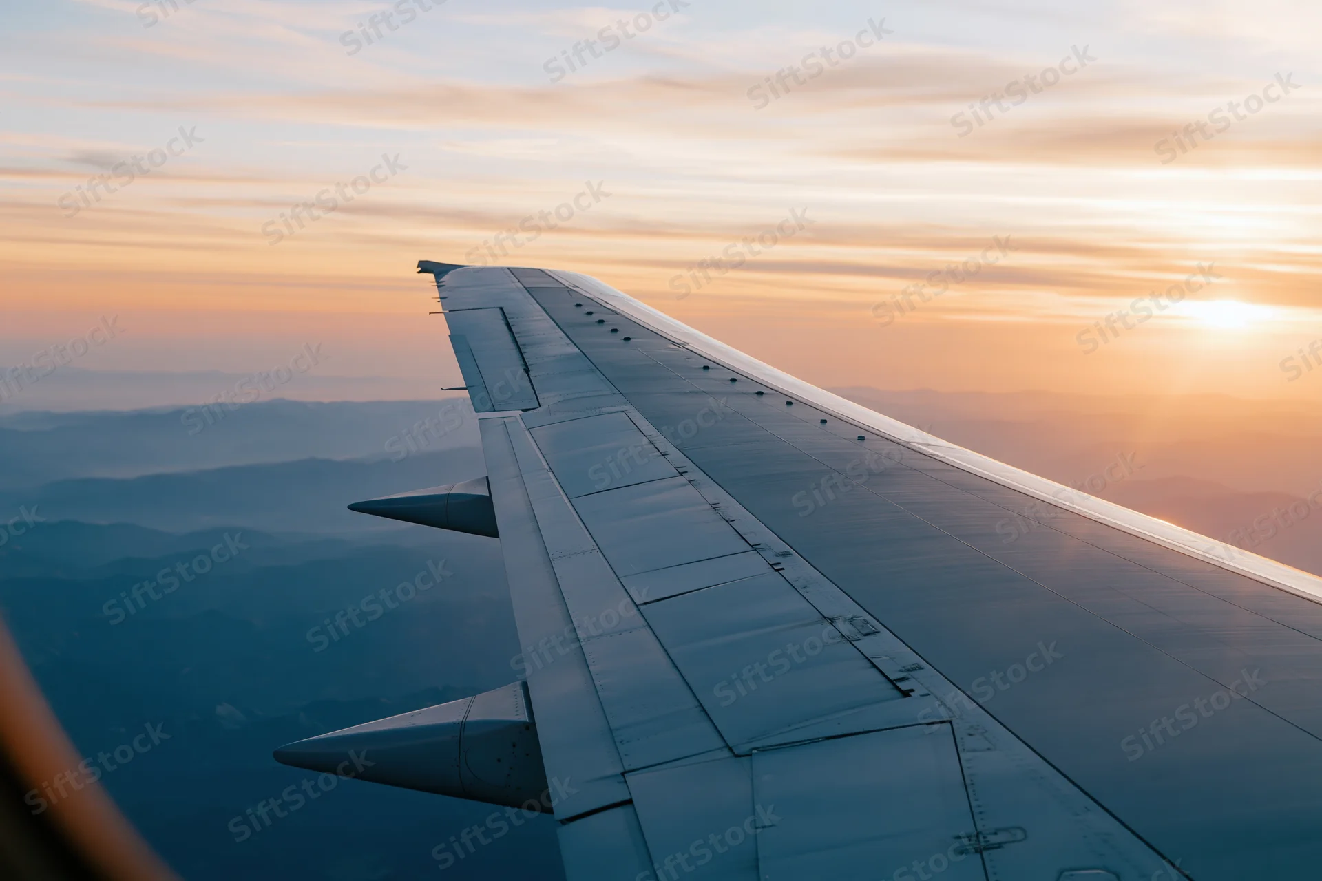 Airplane wing viewed through passenger window during cruising flight at sunset over mountainous landscape
