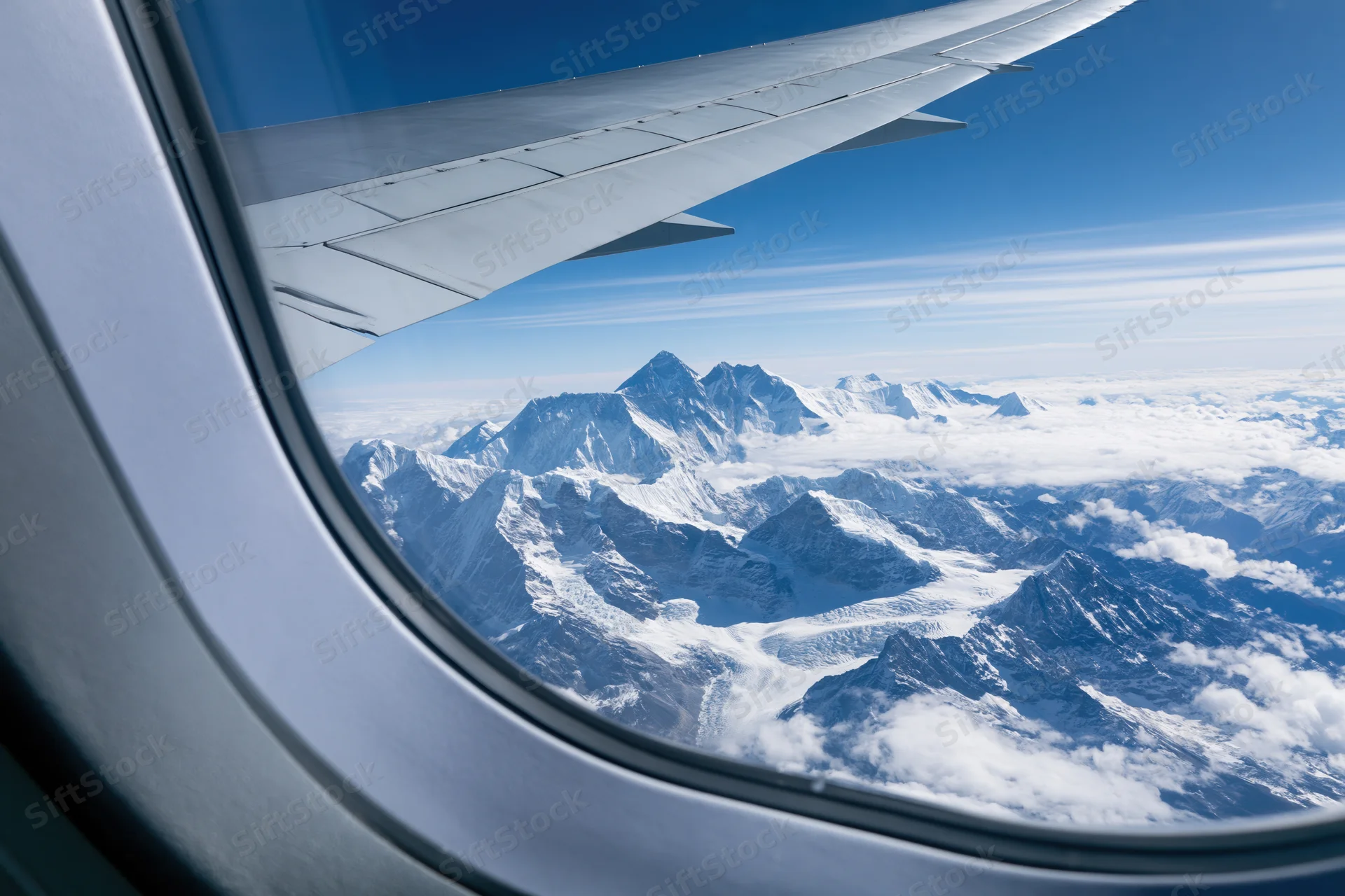Airplane wing visible through window overlooking snow-covered mountain range above clouds