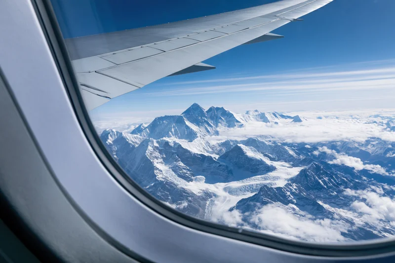 Airplane wing visible through window overlooking snow-covered mountain range above clouds