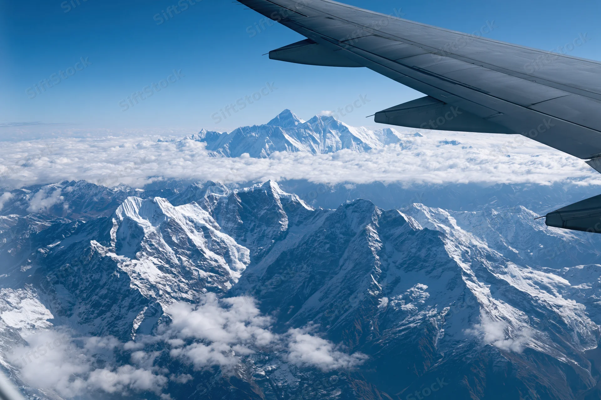 Airplane wing over snow-covered mountain range with clouds below under clear blue sky