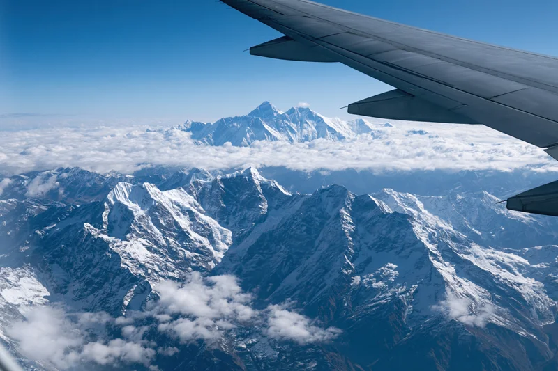 Airplane wing over snow-covered mountain range with clouds below under clear blue sky