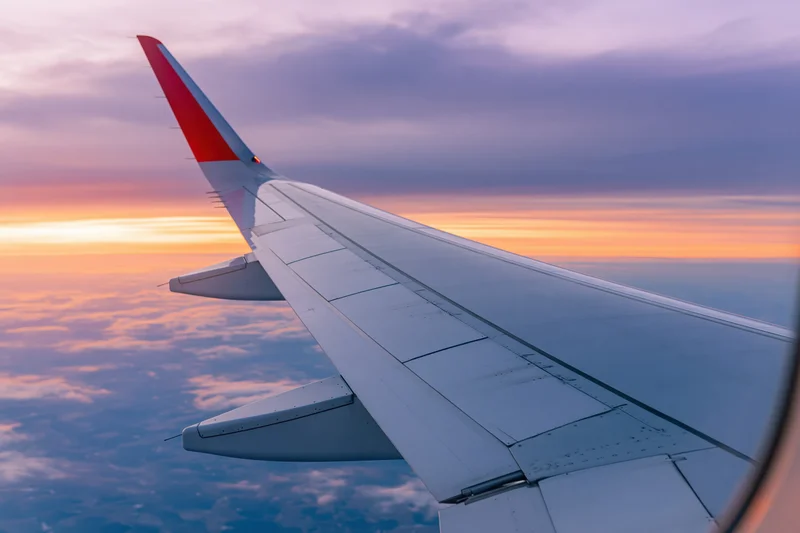 Airplane wing with red tip seen through passenger window during sunset flight above clouds